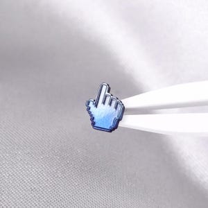May include: A small, metallic blue pin shaped like a computer mouse cursor hand icon. The pin features a gradient finish transitioning from a deep navy blue at the base to a lighter, vibrant blue at the top, with a distinct pixelated edge design.