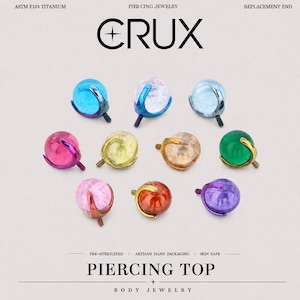 Può includere: Una collezione di nove terminali per piercing sferici con perni filettati, ciascuno con una pietra preziosa colorata tenuta da un'incastonatura a griffe in metallo. Il testo CRUX PIERCING TOP e ASTM F136 TITANIUM REPLACEMENT END è visibile.
