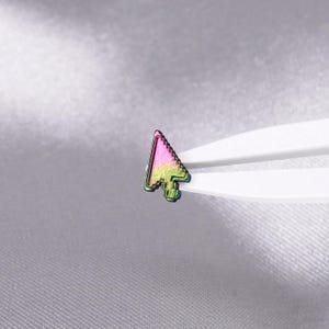 May include: A small, iridescent computer mouse cursor icon pin featuring a gradient of vibrant pink and lime green colors. The metallic cursor shape is held by white plastic tweezers, showcasing its distinct pixelated edge design and reflective finish.