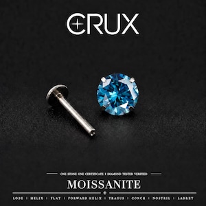 Può includere: Un orecchino a lobo in moissanite blu con perno a base piatta. Il testo CRUX è in alto. Sotto l'orecchino, il testo recita: ONE STONE ONE CERTIFICATE | DIAMOND TESTER VERIFIED, MOISSANITE, e LOBE | HELIX | FLAT | FORWARD HELIX | TRAGUS | CONCH | NOSTRIL | LABRET.