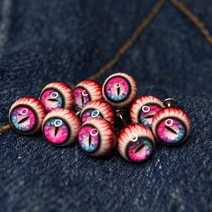 Könnte beinhalten: Eine Ansammlung runder Cabochons mit einem realistischen Augendesign und einer vertikalen Pupille. Die Iris zeigt einen lebendigen Farbverlauf aus Pink-, Lila- und Blautönen, umgeben von einer weißen Sklera mit feinen roten Blutgefäßdetails.