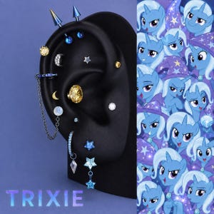 May include: A black ear display model featuring a collection of blue, silver, and gold-toned piercings. The set includes spike studs, star-shaped charms, a crescent moon, a large yellow gem, and a chain-linked hoop earring. The word TRIXIE is visible.