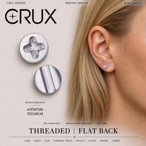 May include: Two silver-colored circular flat back studs with screw-like head designs, one featuring a cross-shaped indentation and the other a single slot. The text CRUX JEWELRY, HYPOALLERGENIC, ASTM F136 TITANIUM, THREADED | FLAT BACK is visible.