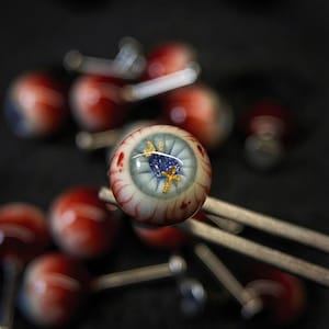 Op de afbeelding: Close-up van verschillende glazen oogcabochons met een religieus thema. Het oog in focus heeft een lichtblauwe iris met een gouden kruis en een blauw hart. De aderen zijn rood en de achtergrond is wazig.