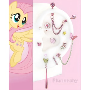 Puede incluir: Una colección de joyas para el cuerpo rosas y plateadas, que incluyen pendientes y una cadena, exhibidas en un modelo de oreja blanca. Las joyas presentan diseños de mariposas, flores y estrellas. La palabra "Fluttershy" está escrita en rosa.