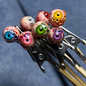 Puede incluir: Una colección de cuentas artesanales de vidrio con forma de globo ocular, con varios colores de iris, incluyendo azul, verde, amarillo y morado. Las cuentas están unidas a pequeños palitos de madera con pinzas metálicas.