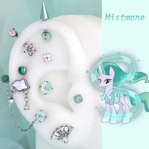 Könnte beinhalten: Eine Sammlung von Ohrpiercings, darunter Stecker, ein Barbell und ein Kettenohrring, die auf einem weißen Ohrmodell präsentiert werden. Die Piercings weisen verschiedene Edelsteine und Designs auf. Das Bild enthält auch eine Cartoon-Illustration eines Einhorns mit dem Text "Mistmane".
