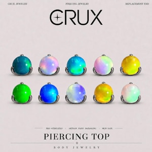 May include: Ten spherical piercing tops arranged in two rows, each featuring a smooth, iridescent, or solid-coloured stone set in a dark metal base. Text reads: CRUX JEWELRY PIERCING JEWELRY REPLACEMENT END, PRE-STERILIZED, ARTISAN HAND PACKAGING, SKIN SAFE, PIERCING TOP, BODY JEWELRY.