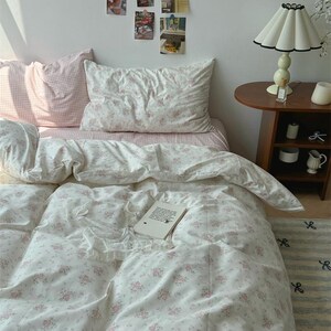 May include: A bed dressed with a floral duvet cover and pillowcases in white and pink. A small side table with a lamp and decorative items is beside the bed. A book rests on the duvet.