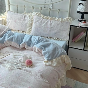 May include: A bed with a white metal frame, adorned with a light pink floral patterned comforter and pillows. The bedding features ruffled edges in a cream color, with a light blue blanket. A small lamp and nightstand are visible.