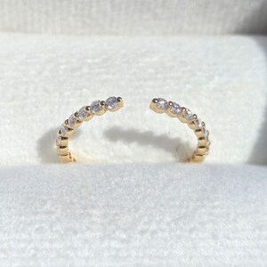 May include: A gold-colored open ring with a row of small, clear gemstones. The ring is displayed in a white velvet jewelry box with a blue lid. The gemstones are evenly spaced along the ring's band.