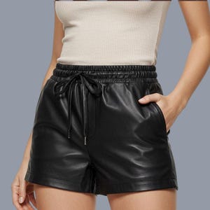 Può includere: Pantaloncini neri in similpelle con elastico in vita e chiusura con coulisse. I pantaloncini hanno due tasche anteriori e una vestibilità rilassata. Il materiale ha una leggera lucentezza. I pantaloncini sono indossati da una persona.