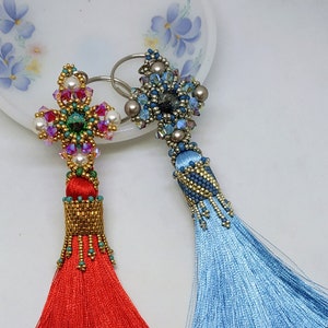 Op de afbeelding: Twee kralen sleutelhangers met kwastjes. De ene sleutelhanger heeft een rode kwast en een gouden en groene kralenbloem. De andere sleutelhanger heeft een blauwe kwast en een zilveren en blauwe kralenbloem. De sleutelhangers liggen op een witte achtergrond met een bloemenpatroon.