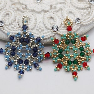 Two handcrafted, intricate beaded snowflake pendants or ornaments, featuring designs in blue/silver/dark blue and green/gold/red/turquoise color palettes, displayed on an ornate white surface.