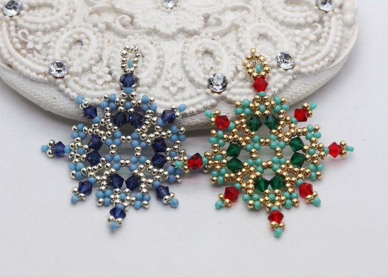 Two handcrafted, intricate beaded snowflake pendants or ornaments, featuring designs in blue/silver/dark blue and green/gold/red/turquoise color palettes, displayed on an ornate white surface.