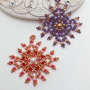 Two handcrafted, intricate beaded snowflake pendants or ornaments displayed on an ornate white lacy surface. The top-right snowflake features purple, gold, and white beads with a central gold pearl, while the bottom-left snowflake features copper, red, and pink beads with a central white pearl.