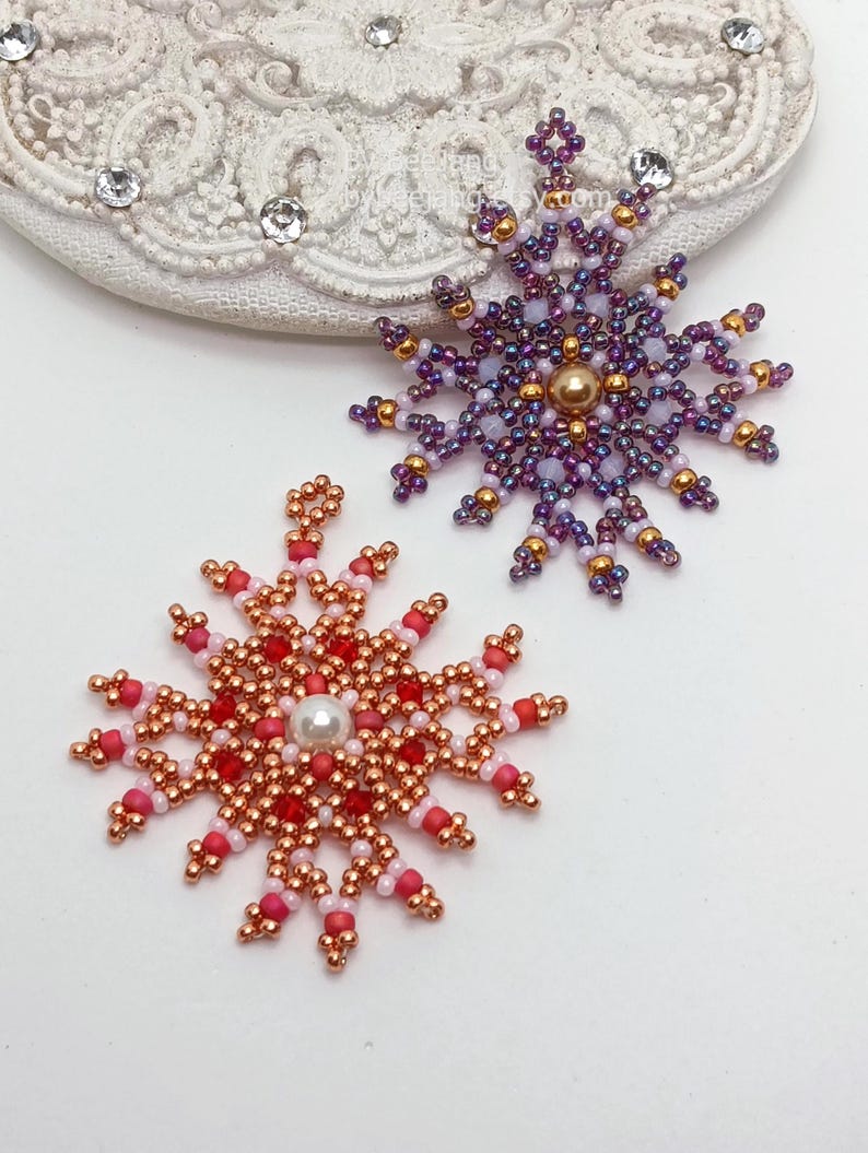 Two handcrafted, intricate beaded snowflake pendants or ornaments displayed on an ornate white lacy surface. The top-right snowflake features purple, gold, and white beads with a central gold pearl, while the bottom-left snowflake features copper, red, and pink beads with a central white pearl.