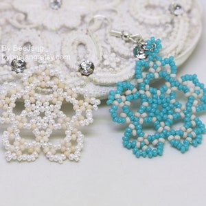 A pair of intricate, handcrafted jewelry pieces, likely earrings, featuring a snowflake or floral design made of seed beads; one is predominantly white/cream colored and the other is turquoise blue and white, displayed on a white background with lace accents.