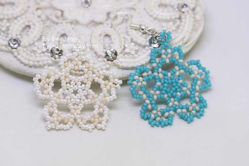 A pair of intricate, handcrafted jewelry pieces, likely earrings, featuring a snowflake or floral design made of seed beads; one is predominantly white/cream colored and the other is turquoise blue and white, displayed on a white background with lace accents.