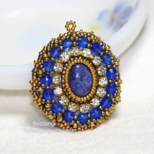 A single, intricate, handcrafted bead-embroidered jewelry piece, likely a pendant or brooch, featuring a central oval blue lapis lazuli stone
surrounded by a border of small, gold-tone seed beads and an outer ring of blue crystal beads and clear rhinestones.