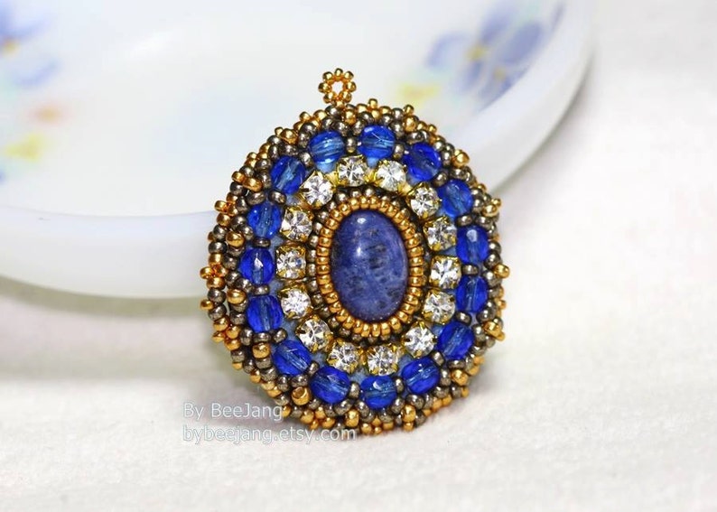 A single, intricate, handcrafted bead-embroidered jewelry piece, likely a pendant or brooch, featuring a central oval blue lapis lazuli stone
surrounded by a border of small, gold-tone seed beads and an outer ring of blue crystal beads and clear rhinestones.