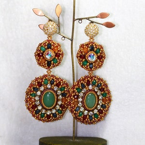A large pair of intricate, handcrafted drop earrings featuring oval green center stones and bead embroidery in gold, red, dark blue, white, green, and peach colors.