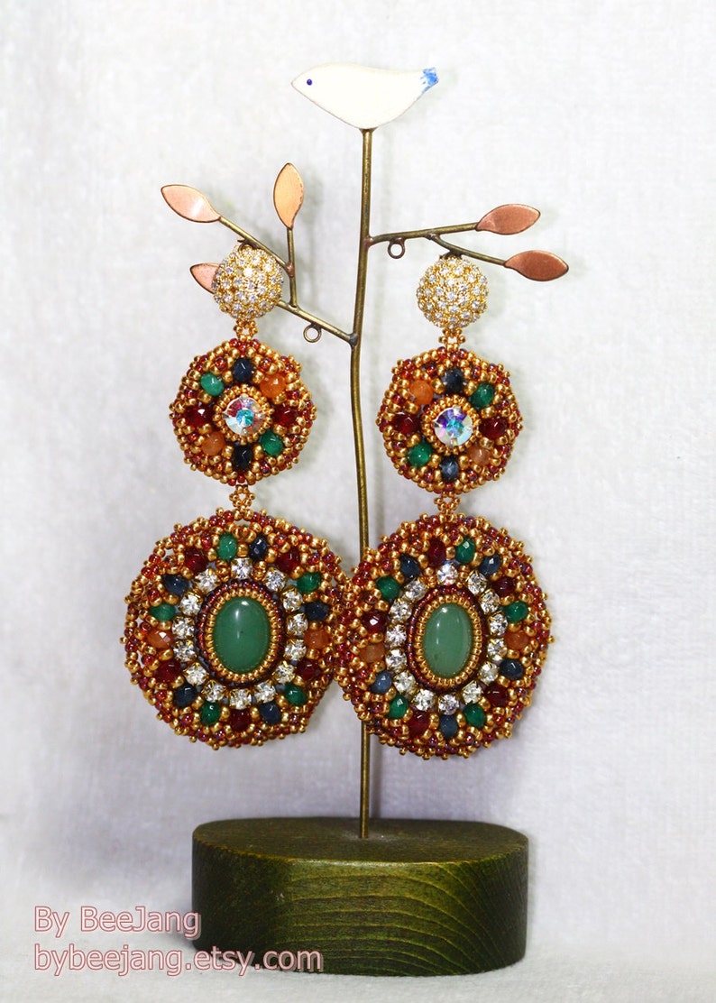 A large pair of intricate, handcrafted drop earrings featuring oval green center stones and bead embroidery in gold, red, dark blue, white, green, and peach colors.