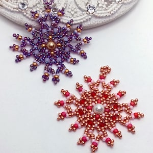 Two handcrafted, intricate beaded snowflake pendants or ornaments displayed on an ornate white surface. The left snowflake features purple, lavender, and gold beads with a central gold faux pearl, while the right snowflake features red, pink, and copper beads with a central white faux pearl.