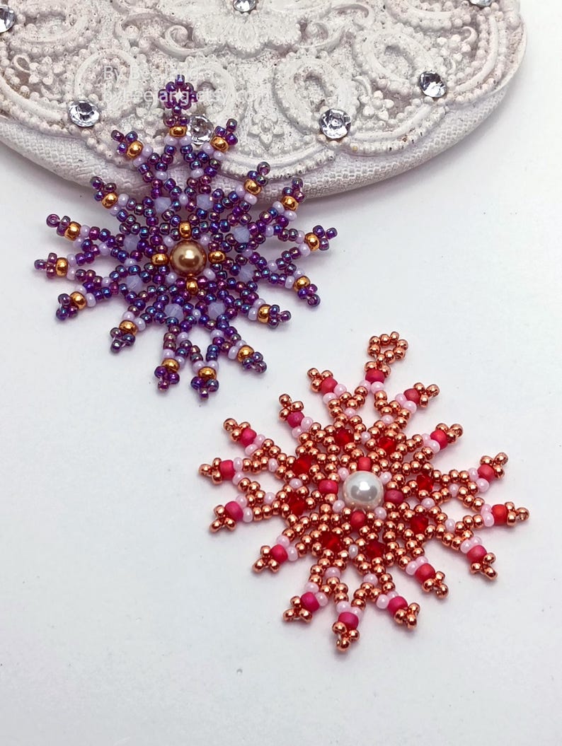 Two handcrafted, intricate beaded snowflake pendants or ornaments displayed on an ornate white surface. The left snowflake features purple, lavender, and gold beads with a central gold faux pearl, while the right snowflake features red, pink, and copper beads with a central white faux pearl.
