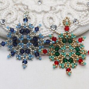 Two handcrafted, intricate beaded snowflake pendants or ornaments, featuring designs in blue/silver/dark blue and green/gold/red/turquoise color palettes, displayed on an ornate white surface.