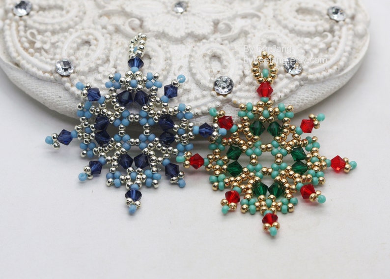 Two handcrafted, intricate beaded snowflake pendants or ornaments, featuring designs in blue/silver/dark blue and green/gold/red/turquoise color palettes, displayed on an ornate white surface.