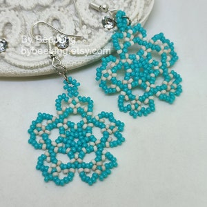 A pair of intricate, handcrafted drop earrings featuring turquoise blue and white seed beads in a snowflake or floral mandala pattern, displayed on a white lace background with small rhinestone details.