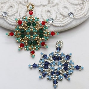 Two handcrafted, intricate beaded snowflake pendants or ornaments, featuring designs in blue/silver/dark blue and green/gold/red/turquoise color palettes, displayed on an ornate white surface.