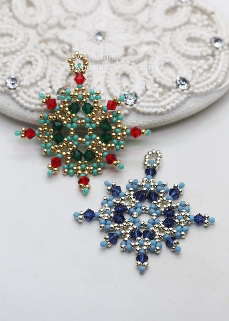 Two handcrafted, intricate beaded snowflake pendants or ornaments, featuring designs in blue/silver/dark blue and green/gold/red/turquoise color palettes, displayed on an ornate white surface.