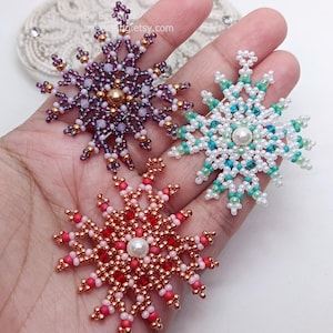 Three handcrafted, intricate beaded snowflake pendants or ornaments being held in a person's hand, featuring detailed designs in purple, mint green/turquoise, and red color palettes.