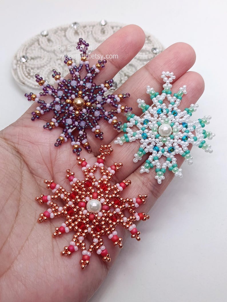Three handcrafted, intricate beaded snowflake pendants or ornaments being held in a person's hand, featuring detailed designs in purple, mint green/turquoise, and red color palettes.