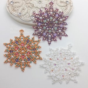 Könnte beinhalten: Drei bestickte Schneeflocken-Ornamente in Lila-, Weiß- und Kupfertönen. Die Schneeflocken sind aufwendig mit Perlen und Kristallen gestaltet. Im Hintergrund befindet sich ein weißes, ovales Dekorationsteil mit Strasssteinen. Der Etsy-Shop-Name ist sichtbar.