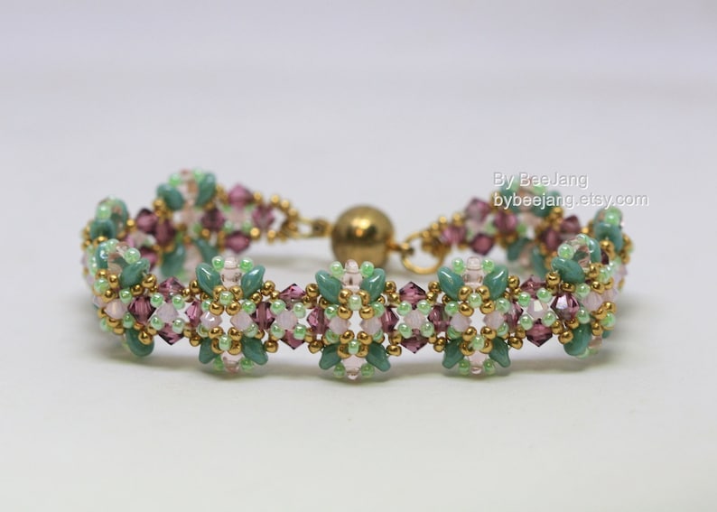 May include: A beaded bracelet featuring a floral design with green, pink, and purple beads. The bracelet has a gold-colored clasp and accents. The text "By BeeJang bybeejang.etsy.com" is visible on the right side.