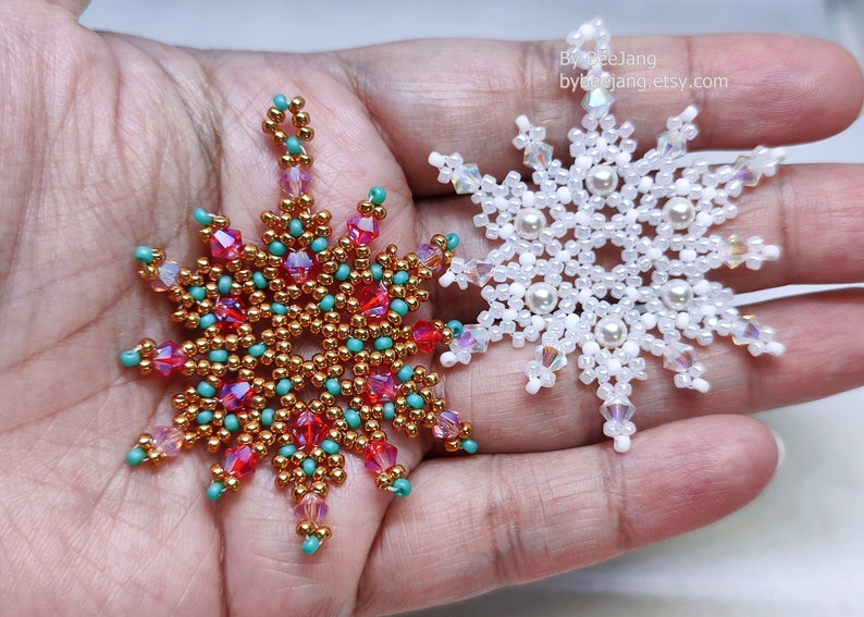 Two handcrafted, intricate beaded snowflake pendants or ornaments being held in a person's hand. The right snowflake features white, clear, and cream faux pearl beads, while the left snowflake features gold, red, turquoise, and pink beads.