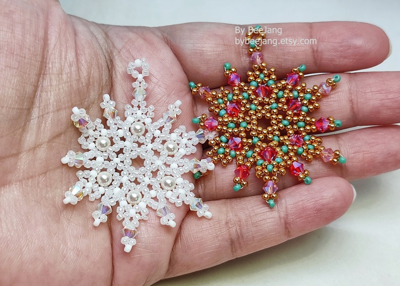 Two handcrafted, intricate beaded snowflake pendants or ornaments being held in a person's hand. The left snowflake features white, clear, and cream faux pearl beads, while the right snowflake features gold, red, turquoise, and pink beads.