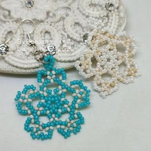 A pair of intricate, handcrafted jewelry pieces, likely earrings, featuring a snowflake or floral design made of seed beads; one is predominantly white/cream colored and the other is turquoise blue and white, displayed on a white background with lace accents.