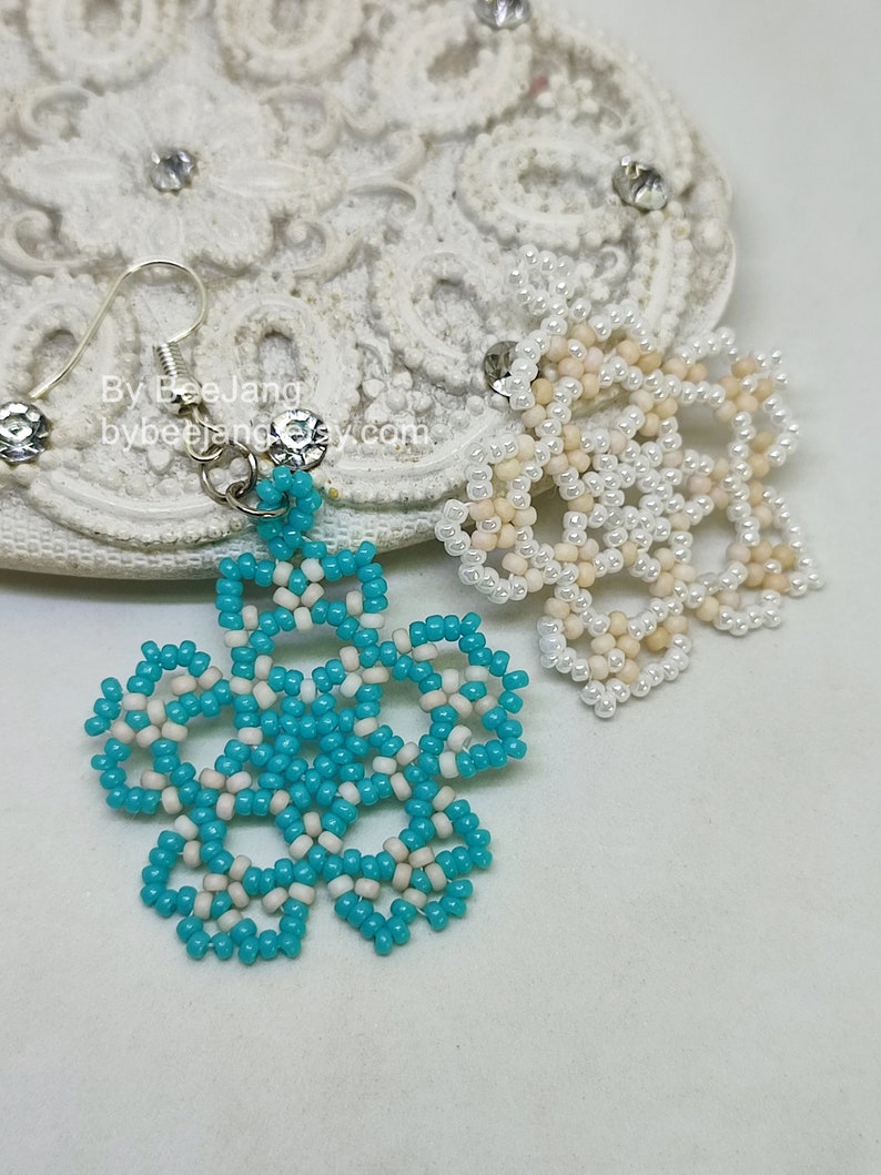 A pair of intricate, handcrafted jewelry pieces, likely earrings, featuring a snowflake or floral design made of seed beads; one is predominantly white/cream colored and the other is turquoise blue and white, displayed on a white background with lace accents.