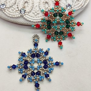 Two handcrafted, intricate beaded snowflake pendants or ornaments, featuring designs in blue/silver/dark blue and green/gold/red/turquoise color palettes, displayed on an ornate white surface.