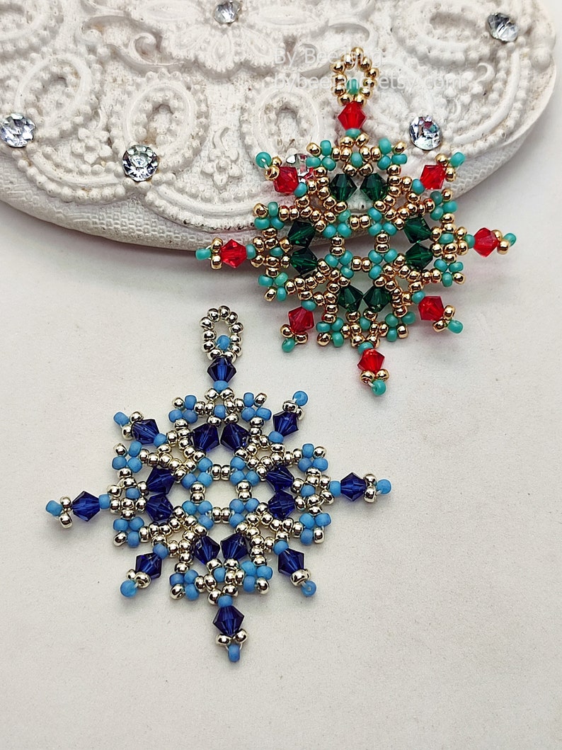 Two handcrafted, intricate beaded snowflake pendants or ornaments, featuring designs in blue/silver/dark blue and green/gold/red/turquoise color palettes, displayed on an ornate white surface.