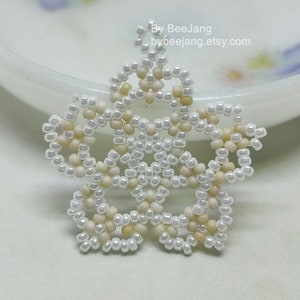 A single, intricate, handcrafted jewelry piece, likely an earring or pendant, featuring white/cream and light yellow seed beads in a star or floral mandala pattern, resting on a white surface.