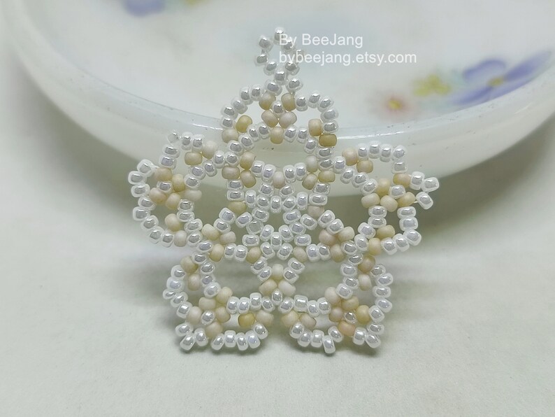 A single, intricate, handcrafted jewelry piece, likely an earring or pendant, featuring white/cream and light yellow seed beads in a star or floral mandala pattern, resting on a white surface.