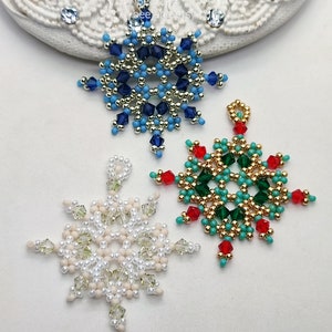 Three handcrafted, intricate beaded snowflake pendants or ornaments, featuring designs in blue/gold/silver, green/gold/turquoise/red, and white/cream/clear color palettes, displayed on an ornate white surface.