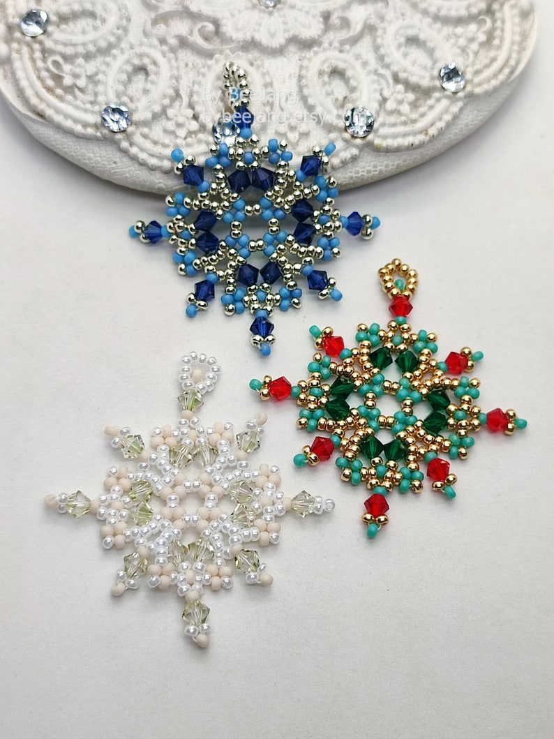 Three handcrafted, intricate beaded snowflake pendants or ornaments, featuring designs in blue/gold/silver, green/gold/turquoise/red, and white/cream/clear color palettes, displayed on an ornate white surface.