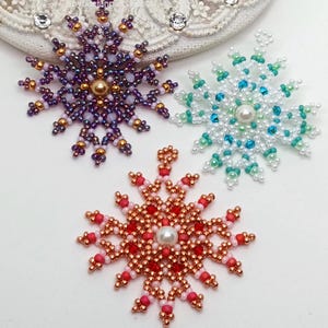Three handcrafted, intricate beaded snowflake pendants or ornaments featuring designs in purple/gold, white/turquoise, and red/copper color palettes, displayed on an ornate white surface.