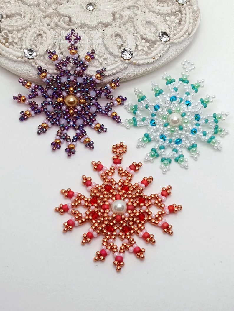 Three handcrafted, intricate beaded snowflake pendants or ornaments featuring designs in purple/gold, white/turquoise, and red/copper color palettes, displayed on an ornate white surface.
